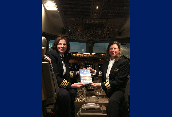 Captains Aileen Watkins (left) and Lynne Rippelmeyer (right) sitting in the cockpit of a Boeing 747 holding the book 747